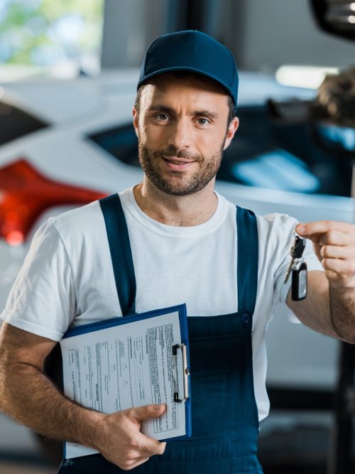 Happy car mechanic looking at camera and holding clipboard and key near car e1653233732554